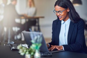 Smiling young African American businesswoman working on a laptop at her desk in a bright modern office with colleagues in the background.